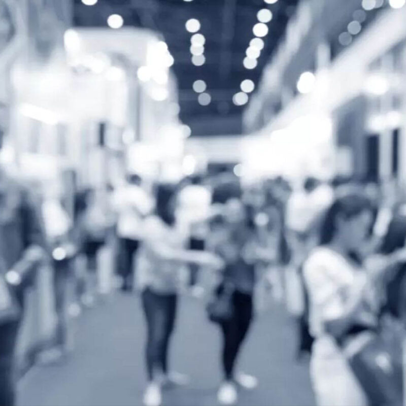 A blurred image of a crowded indoor event with people walking and interacting, showcasing vibrant exhibition booths. The background is filled with bright lights, highlighting the latest in MIM 3D printing technology.