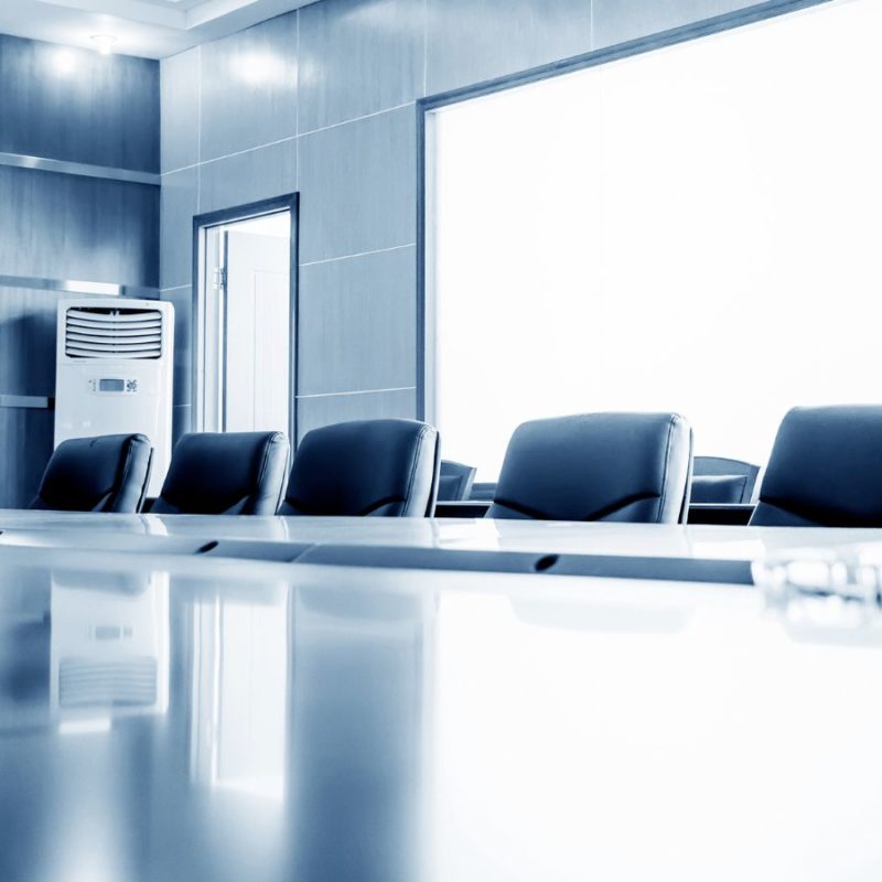 A modern conference room with a long, polished table, black leather chairs, and glass walls, illuminated by bright natural light. An air conditioning unit is visible in the corner.