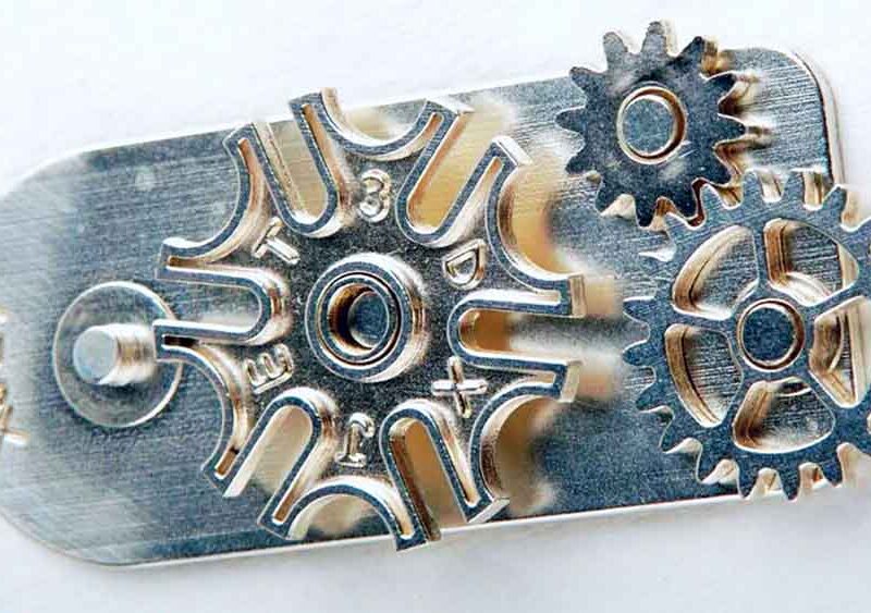 Close-up of a small mechanical assembly with several interlocking gears and a stamped metal plate. One gear is uniquely shaped like a star, and the entire assembly appears metallic and intricate.
