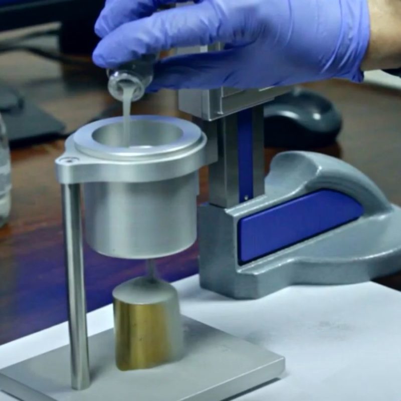 A person wearing blue gloves is pouring liquid from a small bottle into a metal apparatus on a wooden desk with several small bottles in the background.
