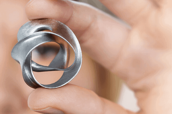 A hand holds a small, abstract, silver metal sculpture with intertwined loops.