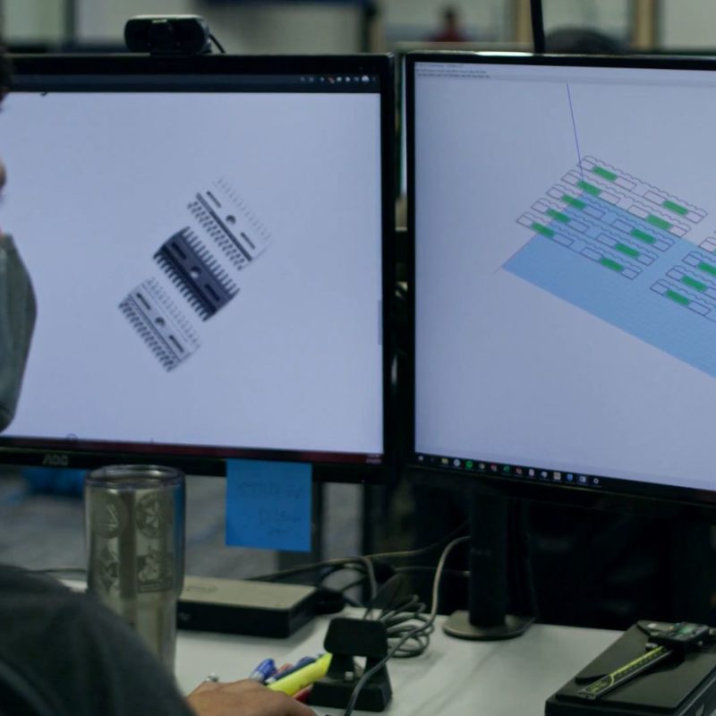 Man wearing a face mask working on two computer monitors displaying technical blueprints in an office setting.