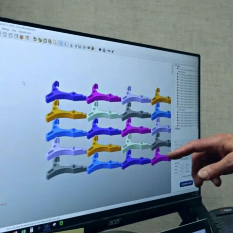 A hand points at a computer screen displaying a 3D modeling software with various colored mechanical parts arranged in a grid.