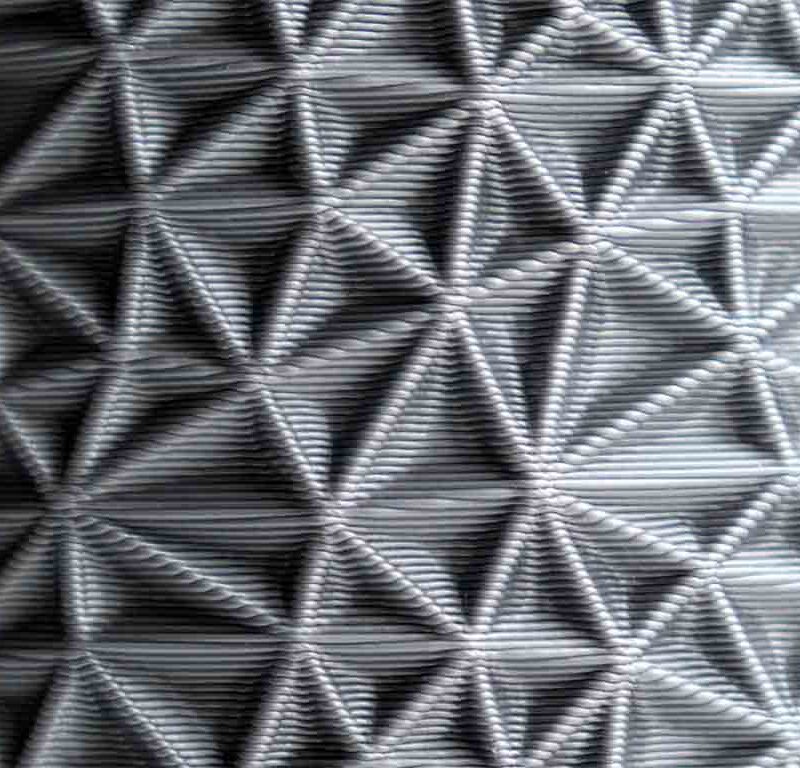 A close-up of a surface with a geometric, triangular pattern, resembling a 3D texture made from interwoven lines. The pattern is repetitive and creates a tactile, structured appearance.