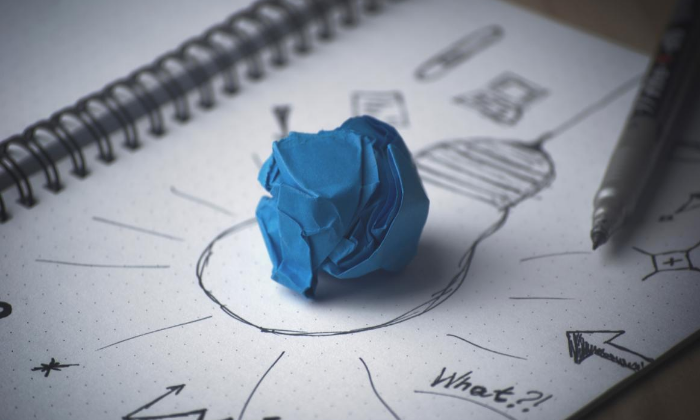 A blue crumpled paper ball on a sketchbook with doodles and a pen nearby, highlighting a creative brainstorming process.