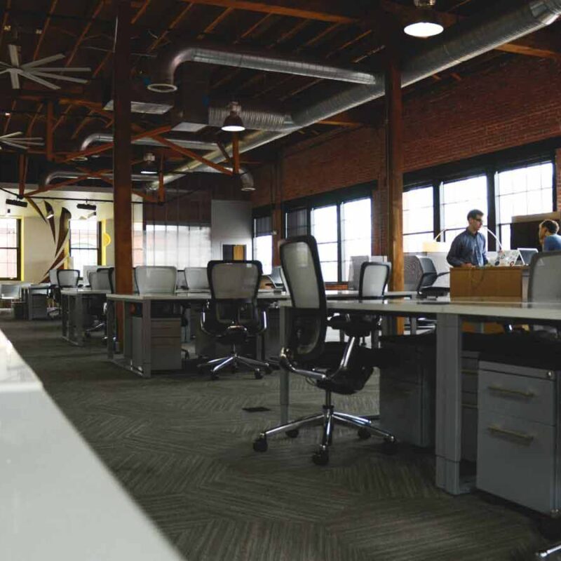 Spacious open office with multiple workstations, ergonomic chairs, industrial design elements, and two people talking in the background near the windows.