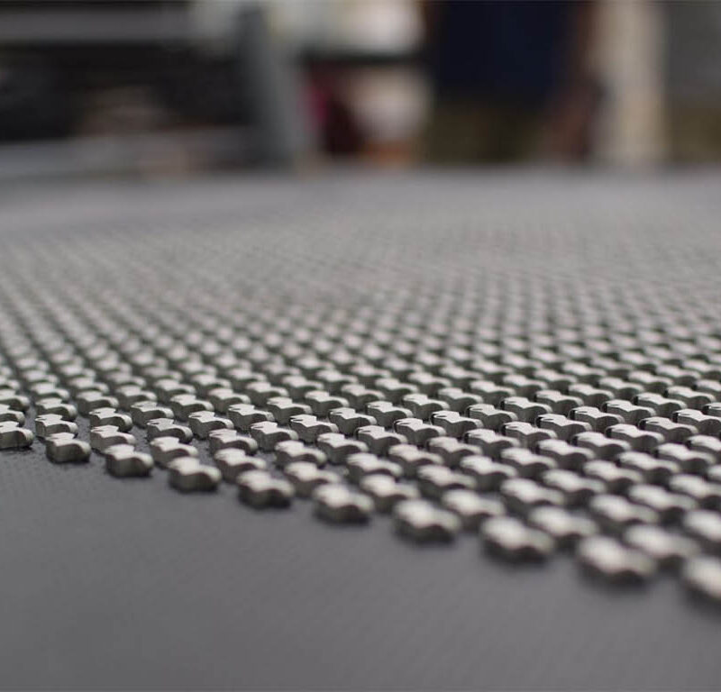 Close-up of a grid-like pattern on a textured surface, with small metallic components arranged uniformly. Blurred background with indistinct shapes and colors.