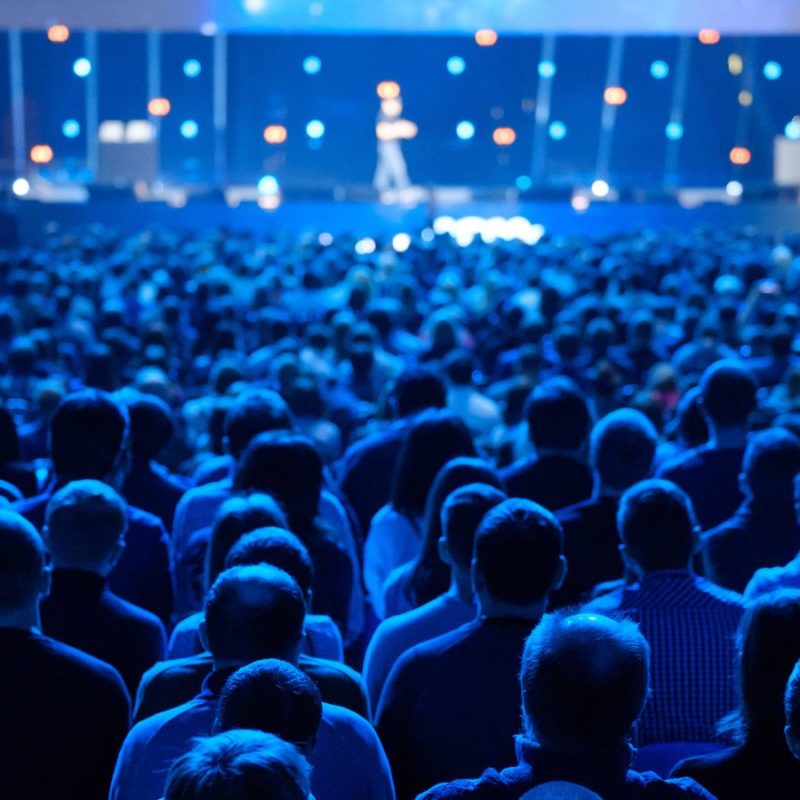 Audience at a conference watching a speaker on stage, illuminated by blue stage lights.