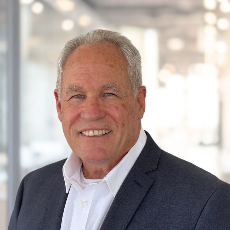 A senior man with a friendly smile, wearing a gray blazer and white shirt, standing in a bright, modern office environment.