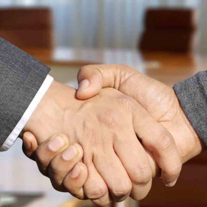 Two individuals shaking hands in a formal setting, with a blurred background of a conference table and chairs.