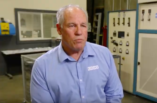 A middle-aged man in a blue shirt with a logo sits beside a 3D metal printer in an industrial setting, looking slightly to the side with a serious expression.