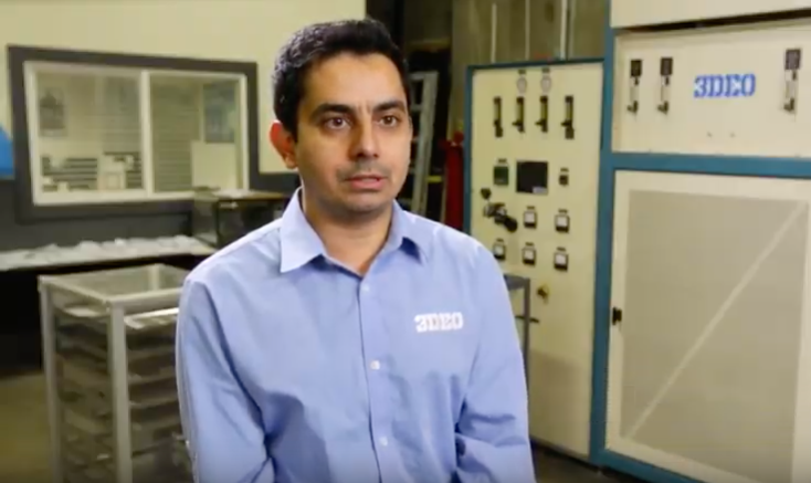 A man in a blue shirt with the logo “3mco” stands in an industrial workspace with a metal 3D printer in the background.