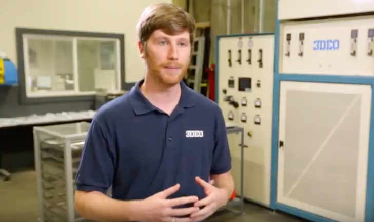 A man in a blue polo shirt standing in an industrial setting with a 3D metal printer in the background.