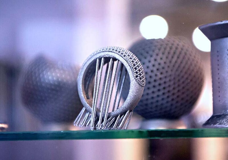 Close-up of intricately designed metallic 3D-printed objects on display, including a sphere with perforations and a ring-like structure with vertical rods. The background is blurred.