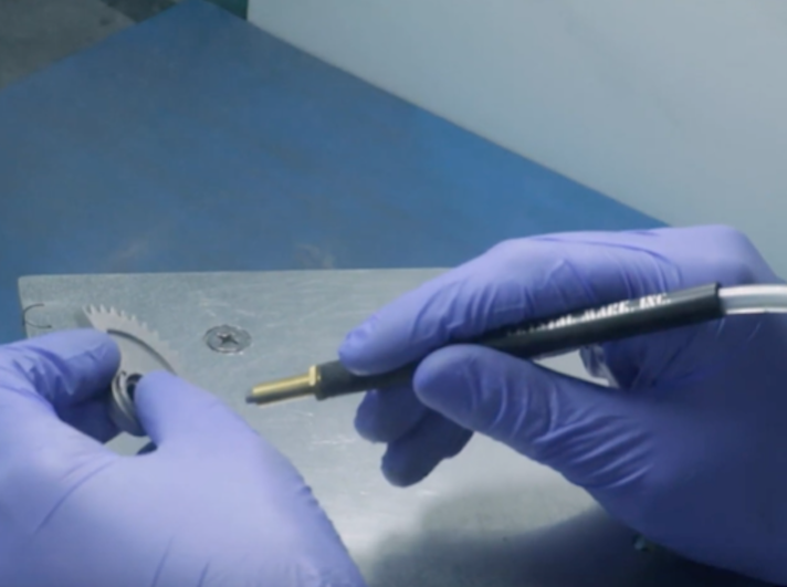 Hands in blue gloves using a precision tool to work on a mechanical part involved in metal 3D printing on a metal surface.