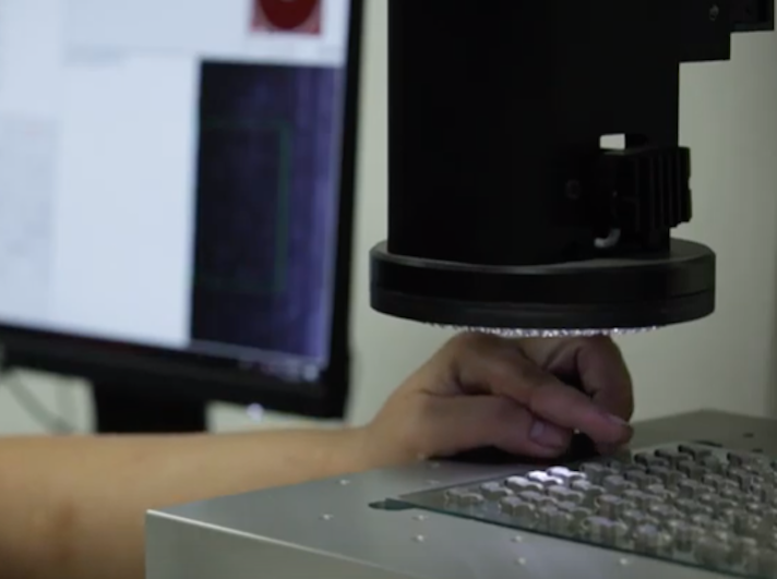 A person's hand operating a keyboard under a large microscope in a laboratory with a 3D metal printer in the background.