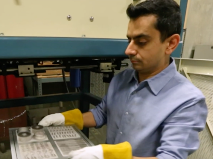 A man inspecting a sheet of metal parts from a 3D metal printer, wearing gloves and a blue shirt.