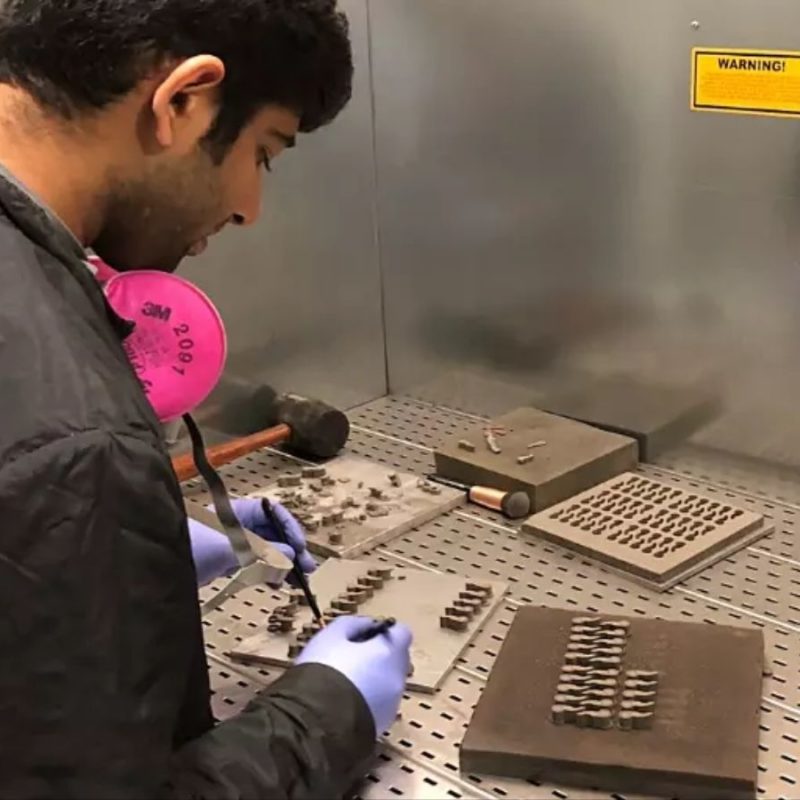 A person wearing a black jacket and blue gloves works on small metallic components inside a lab workstation.