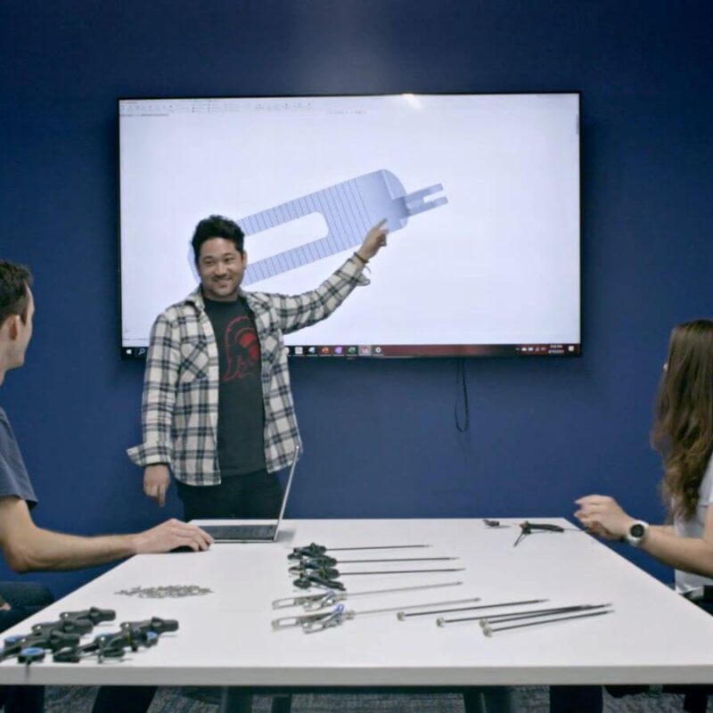 Three people are in a conference room. One person is presenting a CAD drawing of a mechanical part on a wall screen while the other two are seated and listening. Engineering tools are on the table.
