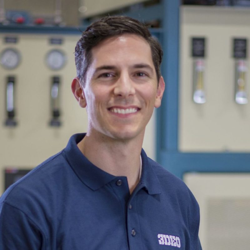 A person wearing a navy-blue collared shirt smiles while standing in front of industrial equipment with gauges and dials.