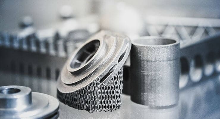 Close-up of several metallic 3D-printed components, including a cylindrical piece and a detailed part with lattice-like structures, on a reflective surface.