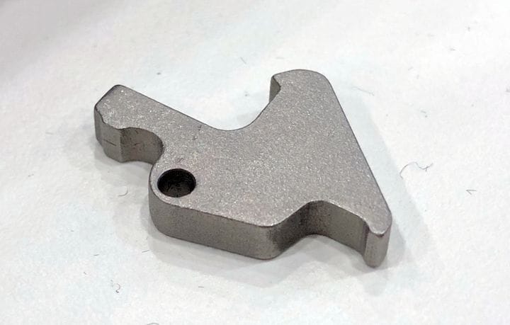 Small metal component with a unique, angular shape and a single hole, resting on a white surface.