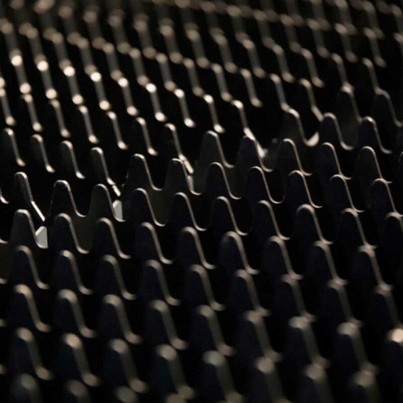 Close-up of a dark textured surface with repetitive, wavy grooves and ridges. The lighting casts subtle shadows, accentuating the undulating pattern.