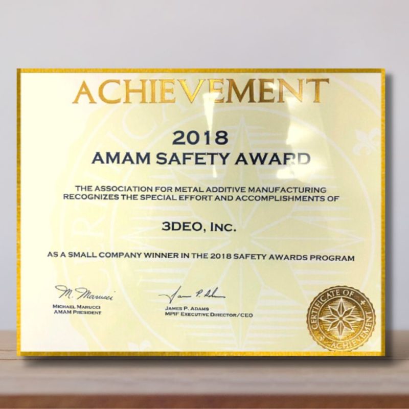 An achievement award certificate for "2018 amam safety award" stands on a wooden surface against a blurred beige background.