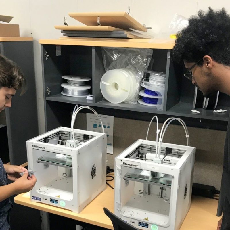Two individuals stand next to two Ultimaker 3D printers in an office, examining them. Shelving units with various supplies are in the background.