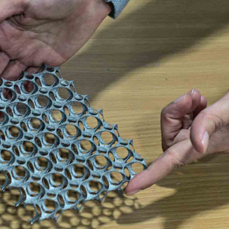 Hands holding and examining a piece of lightweight, flexible, honeycomb-patterned material on a wooden surface.