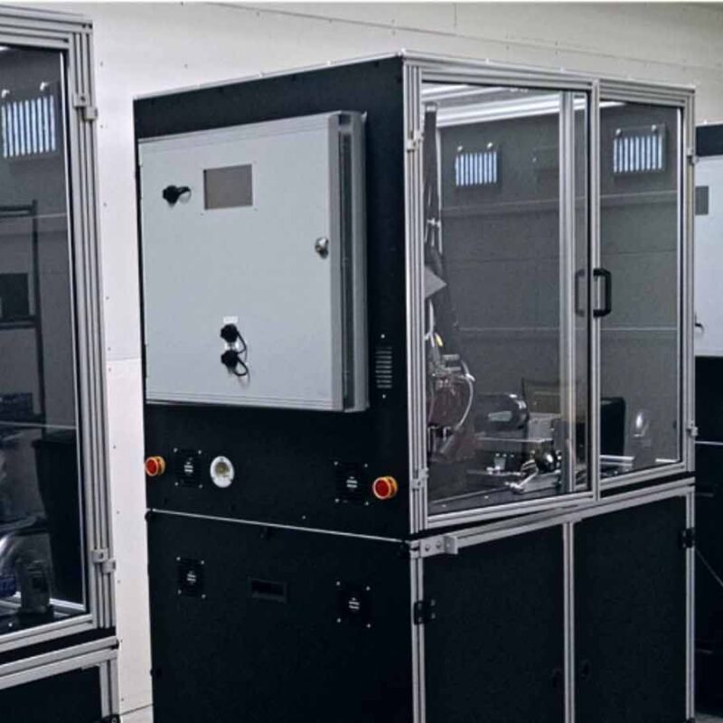 A room with three industrial machines enclosed in glass cabinets, featuring control panels and buttons, placed in a line. Shelves with various items are on the left side of the room.
