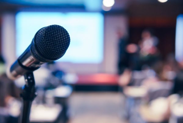 Close-up of a microphone in focus with a blurred background showing a speaker and audience at a metal additive manufacturing conference.