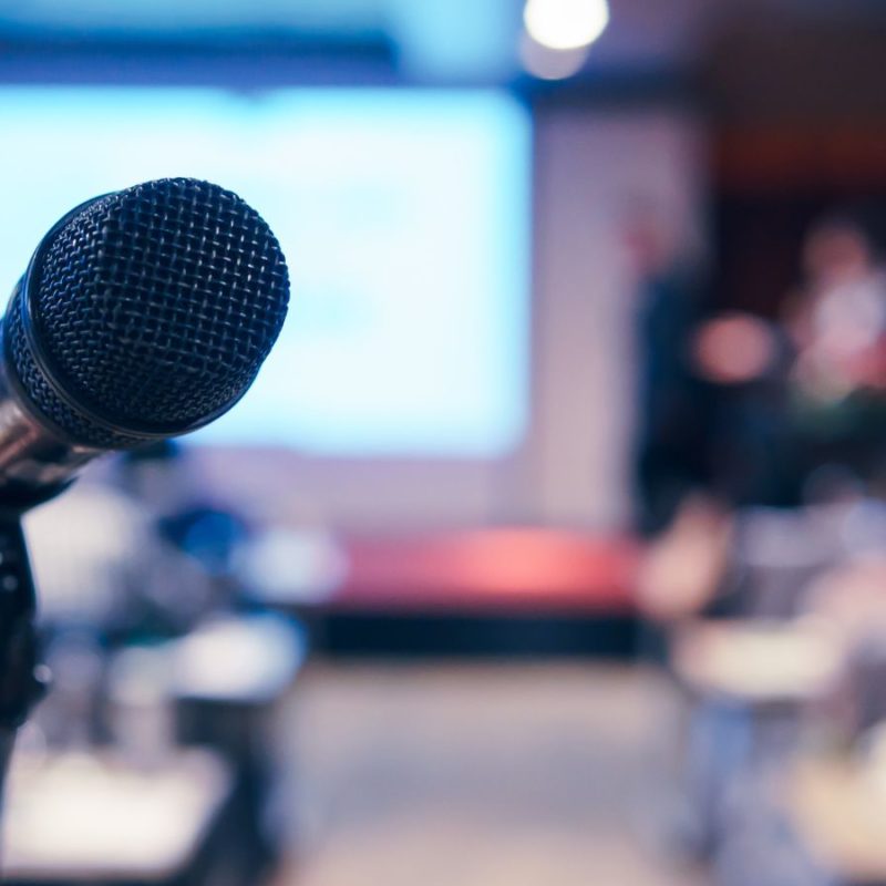 Close-up of a microphone in focus with a blurred background showing a speaker and audience at a metal additive manufacturing conference.