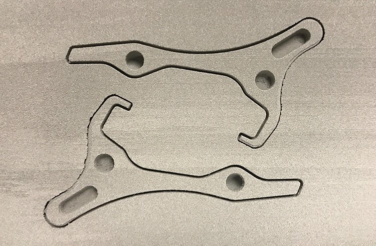 Two dark gray, irregularly shaped metal brackets produced by 3D printing, with holes, lying on a textured light gray surface.