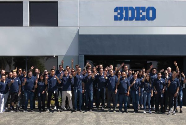 Group of people waving in front of the 3deo metal 3D printing building under a clear sky.