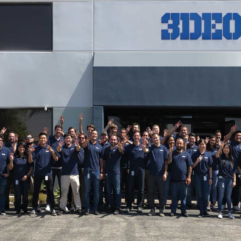 Group of people waving in front of the 3deo metal 3D printing building under a clear sky.