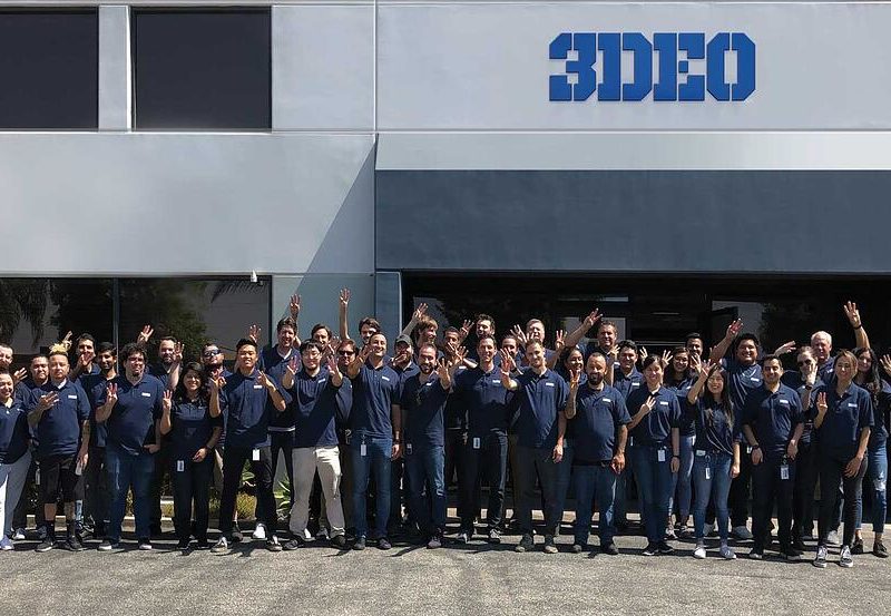 Group of people standing in front of the 3D metal printing building, smiling and waving, dressed in matching blue shirts.