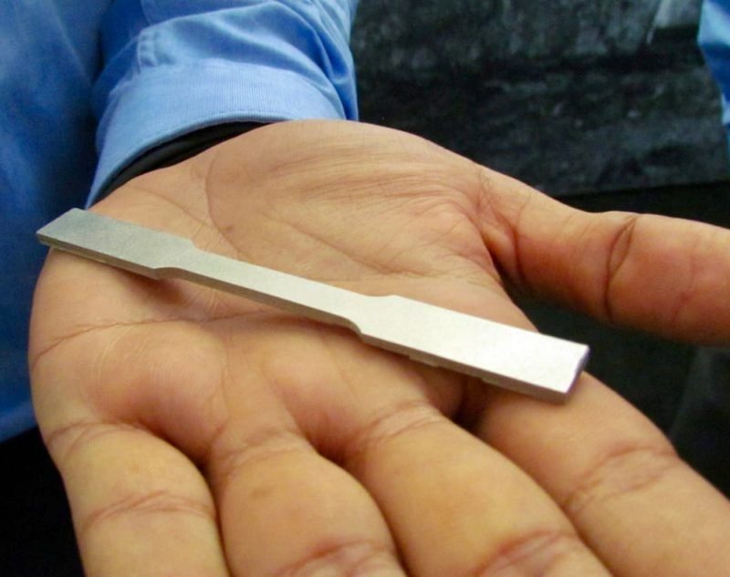 A person holding a metallic key blank produced by a metal 3D printer in their open palm, showcasing it against a blue shirt background.