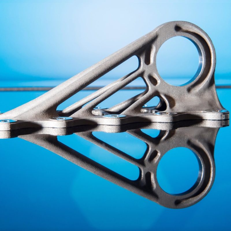 A silver metallic bracket created using 3D metal printing, featuring a complex geometric design, displayed on a reflective blue surface.
