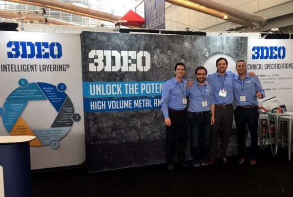 Four men in business attire stand in front of a trade show booth for "3deo," which advertises metal additive manufacturing services.