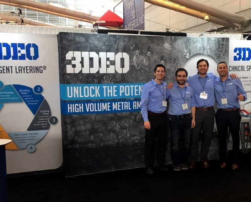 Four men in business attire stand in front of a trade show booth for "3deo," which advertises metal additive manufacturing services.
