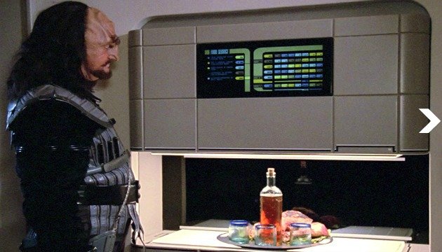 A man in futuristic armor looks at a computer display above a table with food and drinks, showcasing designs from a 3D metal printer.