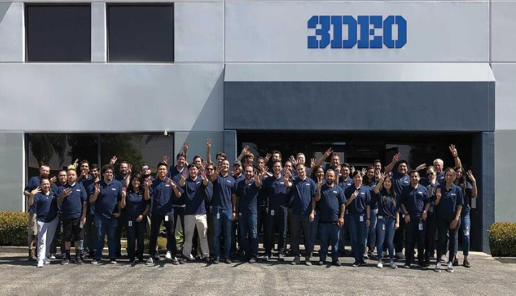 A large group of employees in matching blue shirts posing in front of the "3deo" office building on a sunny day.