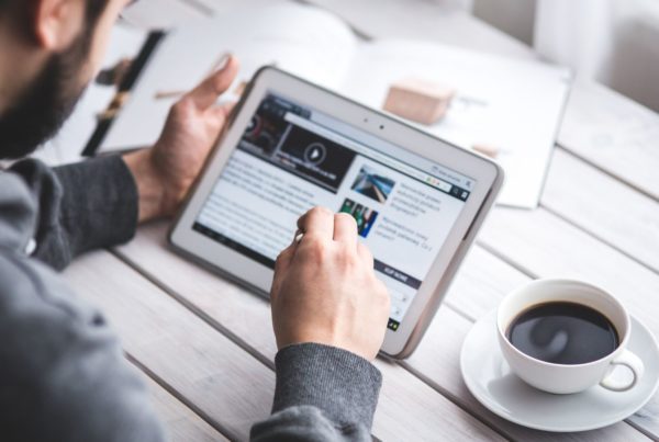 A man browsing news on a tablet with a coffee cup beside him on a white table.