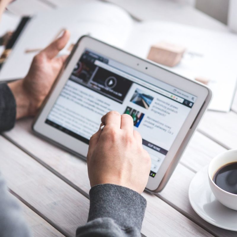 A man browsing news on a tablet with a coffee cup beside him on a white table.