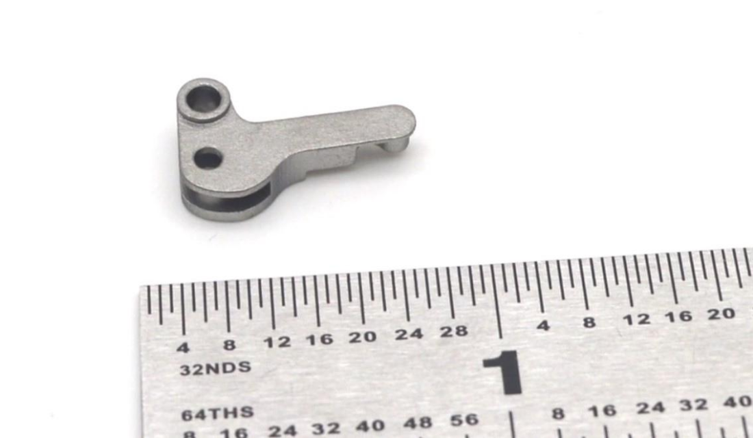A small metal mechanical part with intricate geometries next to a ruler showing measurements in inches.