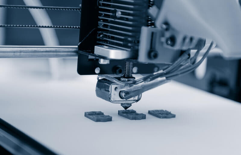 A 3D printer in operation, printing small, intricate objects on a flat surface. The mechanical components and print head are visible in close-up detail.