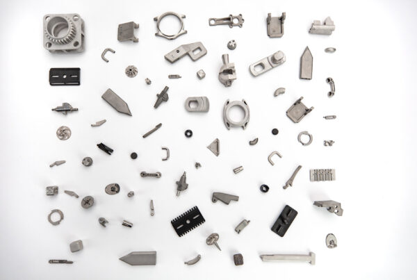 A variety of metal machine parts and components displayed on a white surface, arranged randomly. Items include gears, brackets, and other mechanical elements of different shapes and sizes.