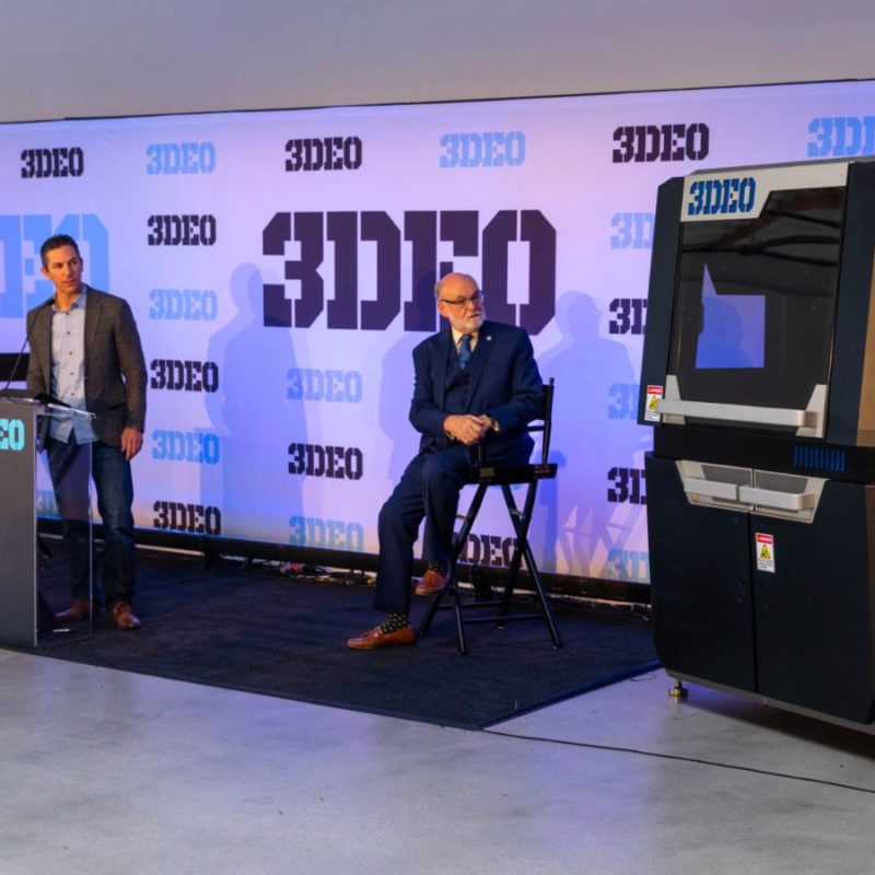 Two men, one standing at a podium and the other seated, at a presentation with a large 3d printer and an audience in a room with a logo-covered backdrop.