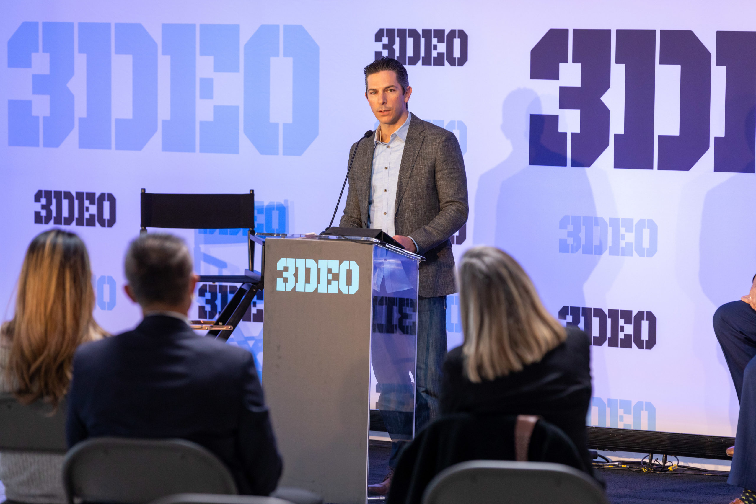 A man stands at a podium labeled "3deo" presenting to an audience in a conference room with "3deo" graphics in the background.